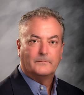 Headshot of a Caucasian man. He has short blonde hair and is wearing a blue collared shirt with a black blazer 